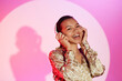 Medium shot of laughing young woman standing at bright pink background looking at camera with hand on headphones listening to upbeat music