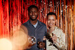 Waist up shot of cheerful Black man holding disco ball while smiling young girl taking his arm, both standing against shining foil background with champagne glasses looking at camera