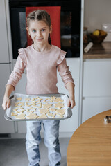 an 8-year-old Caucasian girl holds a baking sheet with raw cookies against the background of an oven in a light kitchen.