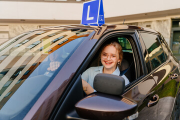 Young positive smiling teen driver with L Plate on the roof of car. Young female try to parking vehicle at city street.