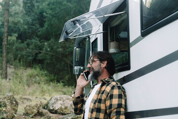 One man outside a motorhome do a phone call with a smile. People and alternative travel van life lifestyle. Alone adult male using cell. Nature park woods in background. Vacation road trip camper