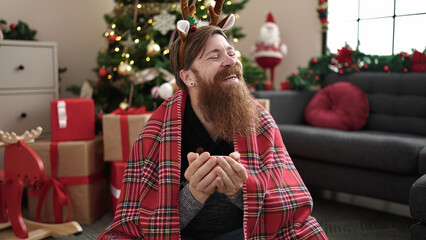 Wall Mural - Young redhead man drinking coffee sitting on floor by christmas tree at home