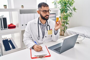 Wall Mural - Young hispanic man doctor holding pills bottle writing on document at clinic