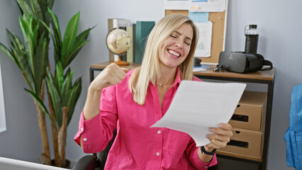 Sticker - A cheerful blonde woman wearing a pink shirt exults while reading a paper in a modern office setting.