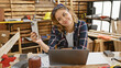 © Krakenimages.com - Young, beautiful hispanic woman carpenter captures her online woodworking business on laptop while taking a smiling selfie at carpentry workshop