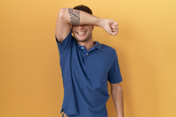 Sticker - Young hispanic man standing over yellow background covering eyes with arm smiling cheerful and funny. blind concept.