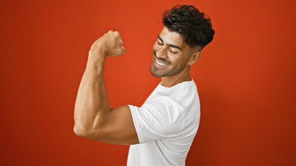 Wall Mural - Handsome young hispanic man flexing muscles with a smile against a red wall background