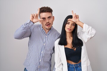 Wall Mural - Young hispanic couple standing over white background making fun of people with fingers on forehead doing loser gesture mocking and insulting.