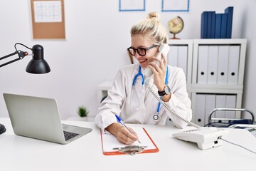 Poster - Young blonde woman doctor talking on telephone writing on document at clinic