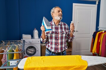 Sticker - Cheerful elder man, with a confident smile, lost in thought while ironing clothes in the laundry room, his grey hair catching the light as he looks away, a portrait of happiness amidst chores.