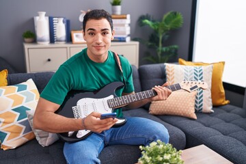 Sticker - Young hispanic man playing electrical guitar using smartphone at home