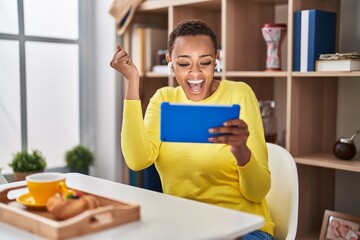 Sticker - African american woman using touchpad sitting on the table at breakfast screaming proud, celebrating victory and success very excited with raised arm