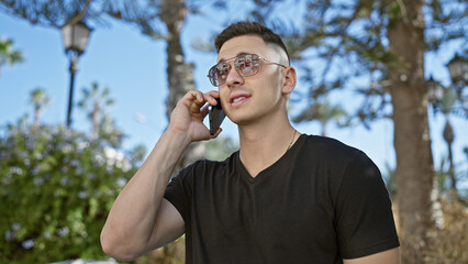 Poster - A young hispanic man in sunglasses talking on a cellphone outdoors in a city park.