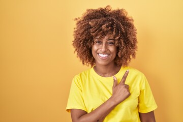 Sticker - Young hispanic woman with curly hair standing over yellow background cheerful with a smile of face pointing with hand and finger up to the side with happy and natural expression on face