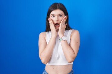 Canvas Print - Young caucasian woman standing over blue background afraid and shocked, surprise and amazed expression with hands on face