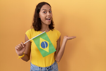 Poster - Young hispanic woman holding brazil flag pointing aside with hands open palms showing copy space, presenting advertisement smiling excited happy