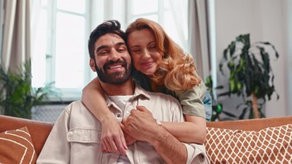 Sticker - Beautiful adult couple hugging, sitting on the sofa by the window in the living room at home, chatting, spending time together romantically.