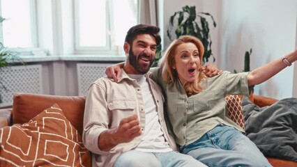 Sticker - Adult couple with remote control watching movie on tv while having fun sitting on sofa near window in living room at home.