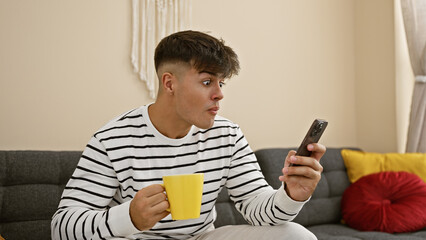 Sticker - Stunned! young hispanic guy's face of surprise while sitting at home, using smartphone, sipping coffee