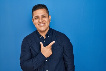 Poster - Hispanic young man standing over blue background cheerful with a smile of face pointing with hand and finger up to the side with happy and natural expression on face