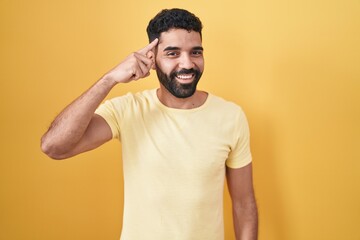 Wall Mural - Hispanic man with beard standing over yellow background smiling pointing to head with one finger, great idea or thought, good memory