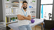 © Krakenimages.com - Young hispanic man with beard smiling confidently while standing arms crossed in a modern rehabilitation clinic interior