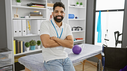 Young hispanic man with beard smiling confidently while standing arms crossed in a modern rehabilitation clinic interior