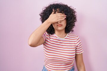 Poster - Young middle east woman standing over pink background covering eyes with hand, looking serious and sad. sightless, hiding and rejection concept