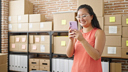 Wall Mural - Young chinese woman ecommerce business worker using smartphone smiling at office