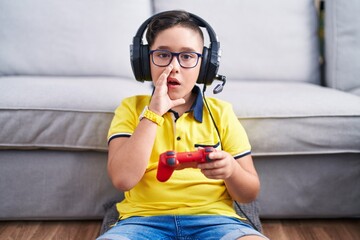 Canvas Print - Young hispanic kid playing video game holding controller wearing headphones hand on mouth telling secret rumor, whispering malicious talk conversation