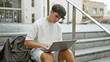 Cool young hispanic teenager, a smart university student engrossed in online study on laptop while sitting casual on stairs at campus, portraying relaxed concentration under city sunlight.