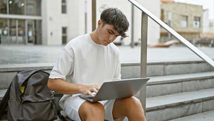 Cool young hispanic teenager, a smart university student engrossed in online study on laptop while sitting casual on stairs at campus, portraying relaxed concentration under city sunlight.