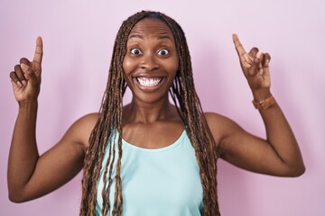 Wall Mural - African american woman standing over pink background smiling amazed and surprised and pointing up with fingers and raised arms.