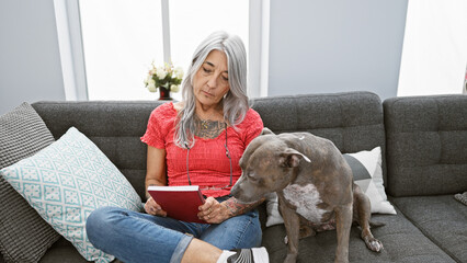 Poster - Relaxed middle age woman, a grey-haired bibliophile, snuggled up with her loyal dog, comfortably sitting on her home sofa, indulging in the leisure of reading a riveting book in her cozy living room.