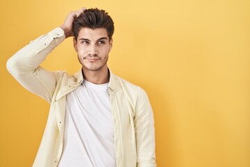 Poster - Young hispanic man standing over yellow background confuse and wondering about question. uncertain with doubt, thinking with hand on head. pensive concept.