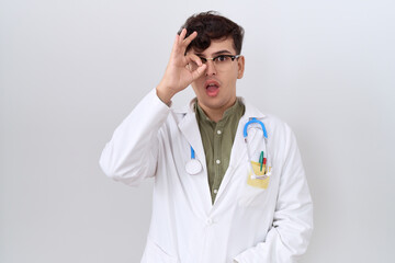 Sticker - Young non binary man wearing doctor uniform and stethoscope doing ok gesture shocked with surprised face, eye looking through fingers. unbelieving expression.