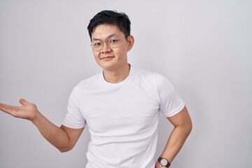 Sticker - Young asian man standing over white background smiling cheerful presenting and pointing with palm of hand looking at the camera.