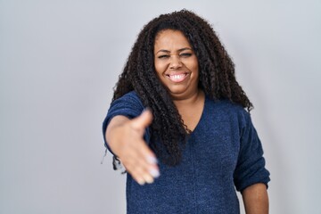 Poster - Plus size hispanic woman standing over white background smiling friendly offering handshake as greeting and welcoming. successful business.