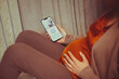 Pregnant woman holding smartphone using pregnancy tracker app on mobile phone while sitting indoors
