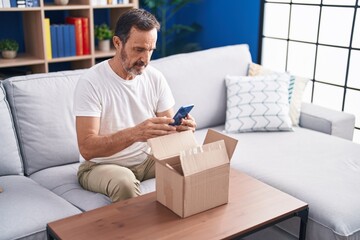 Wall Mural - Middle age man using smartphone unpacking cardboard box at home