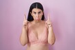 Young hispanic woman wearing pink bra pointing up looking sad and upset, indicating direction with fingers, unhappy and depressed.