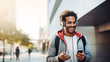 Fototapeta  - Portrait of happy young afro man using his mobile phone with headphones for listening to music outdoors. Technology, urban and lifestyle concept.