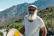 Active cool happy bearded old hipster man standing in nature park holding skateboard. Mature traveler skater enjoying freedom spirit and extreme sports hobby with mountains background
