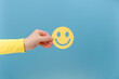 Close up of female hand holding yellow happy smile face, isolated over blue background with copy space. Mental health positive thinking and growth mindset, health care recovery to happiness emotion