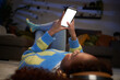 © CarlosBarquero - Unrecognizable teenage girl lying on living room carpet using mobile, blank empty screen. Black gen z woman looking cell phone with headphones at night home. Mock up concept and people domestic life