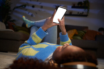 Unrecognizable teenage girl lying on living room carpet using mobile, blank empty screen. Black gen z woman looking cell phone with headphones at night home. Mock up concept and people domestic life