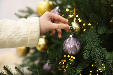 woman decorating fir tree with golden christmas ball indoors, closeup