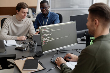 Male programmer writing software code on computer while sitting at desk in IT company office with coworkers