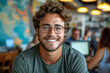 © Gonzalo - Cheerful man with glasses and a green t-shirt is smiling at the camera, seated in a modern office with co-workers and computers in the background.