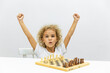 Cute thoughtful girl with blonde curly hair playing chess at table on white background. Early childhood development